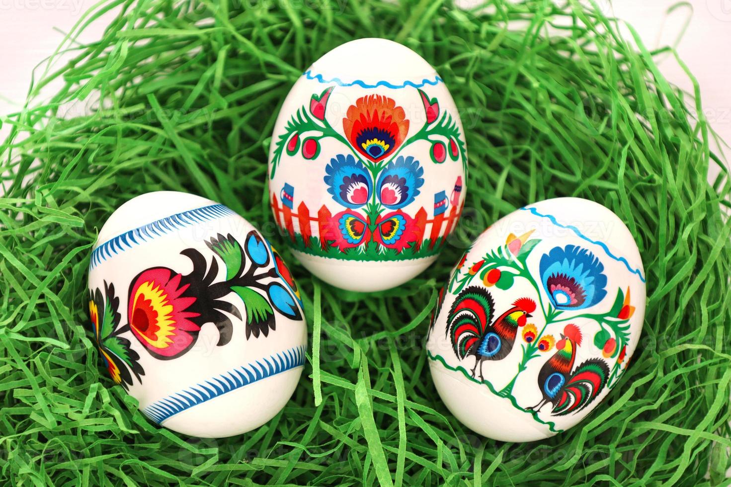 Three Colourful Easter Eggs with floral and roosters folk art pattern printed on stickers. Eggs in green paper grass nest. Polish traditional paper cut from Lowicz region. photo