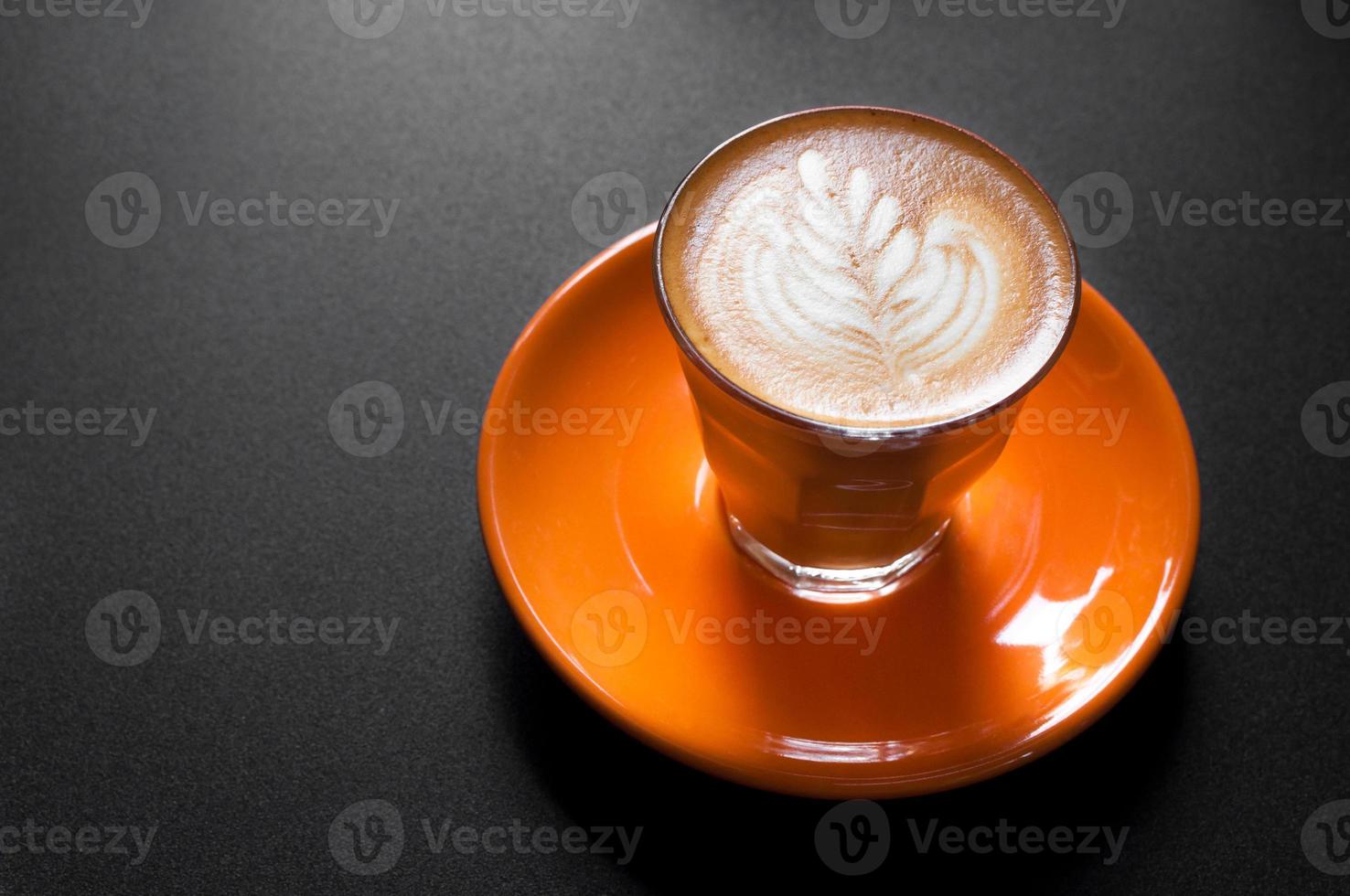Hot latte in orange cup with floral pattern in foam on a dark background photo