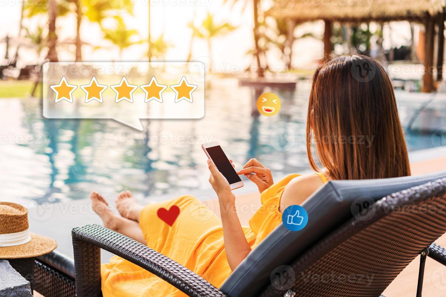 Young woman relaxing at resort pool and using smartphone to give a five-star satisfaction rating of the hotel's service and rooms on social media. Travel lifestyle concept photo