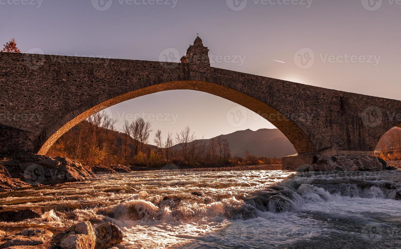 paisaje de un medieval puente terminado un turbulento río a puesta de sol foto