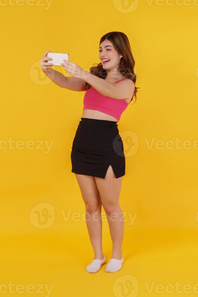 Portrait of Smiling young woman selfie with mobile phone in her hands while standing isolated over yellow background photo