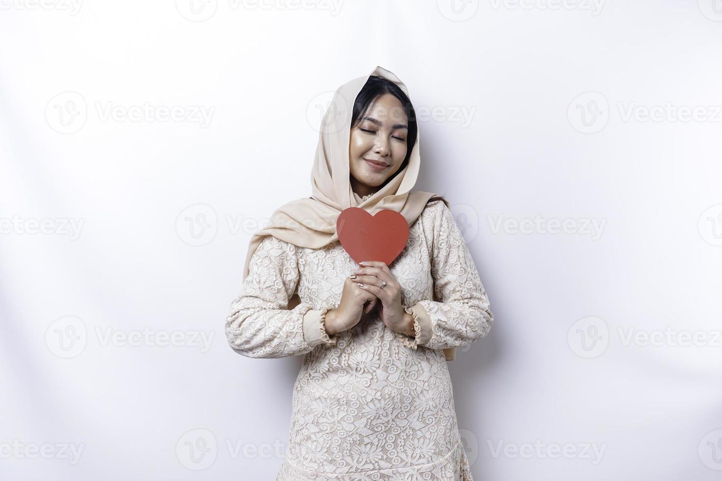 A happy young Asian Muslim woman wearing a hijab feels romantic shapes heart gesture expressing tender feelings and holding a red heart-shaped paper photo