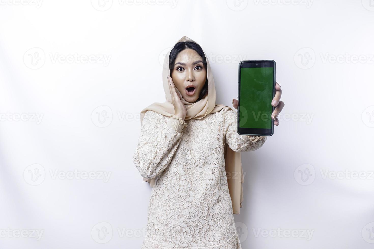 Shocked Asian woman wearing hijab, showing copy space on her phone screen, isolated by white background photo