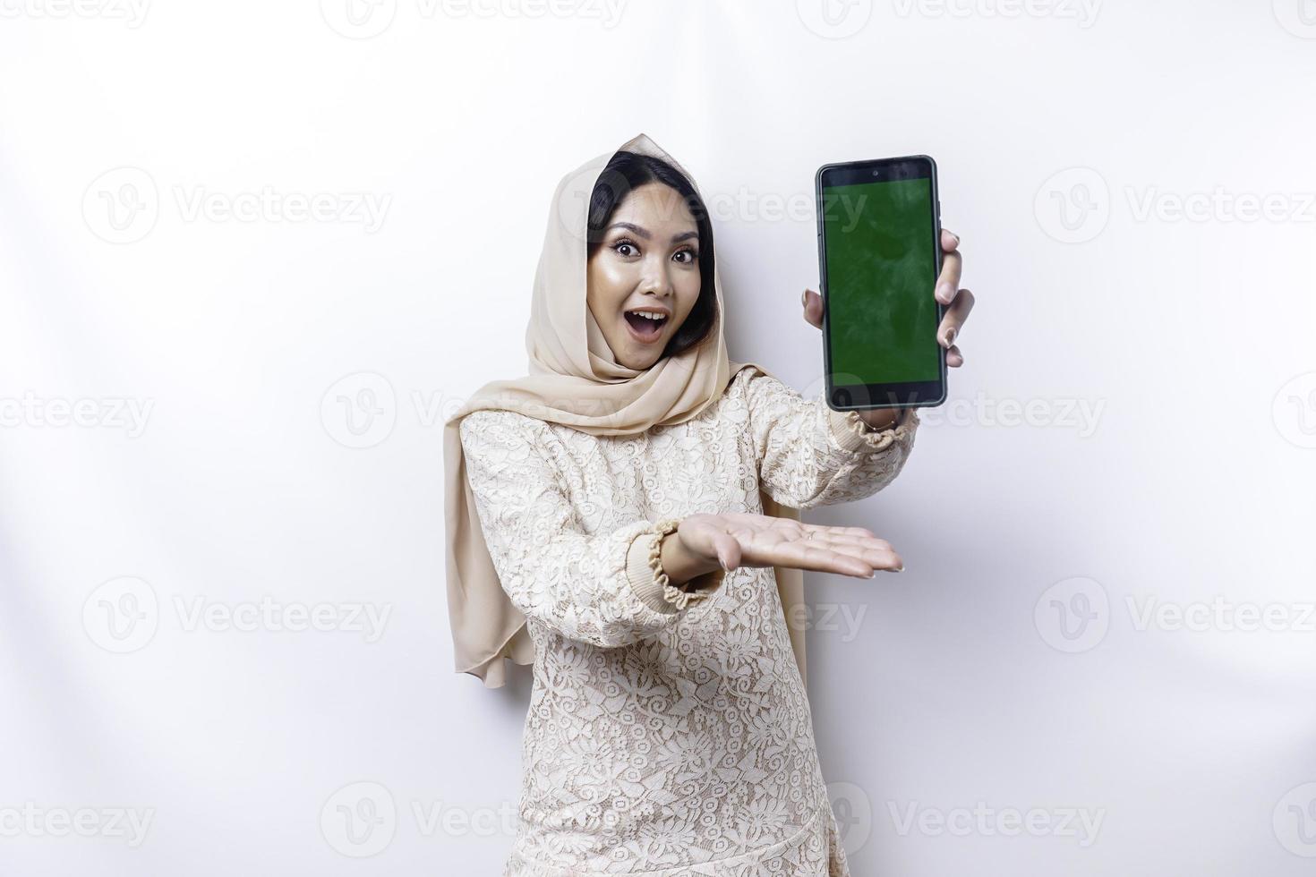A portrait of a happy Asian Muslim woman wearing a hijab, showing her phone screen, isolated by white background photo