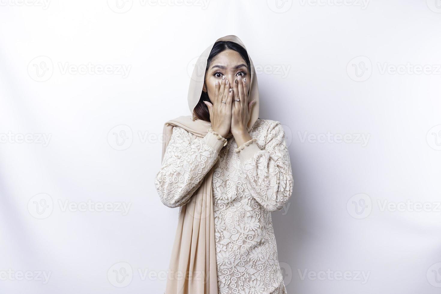 Oops Surprised young Asian Muslim woman covering mouth with hands and staring at camera while standing against white background photo