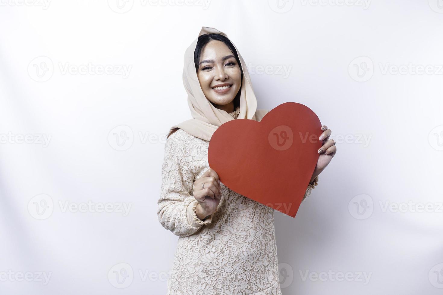 un contento joven asiático musulmán mujer vistiendo un hijab siente romántico formas corazón gesto expresando oferta sentimientos y participación un rojo en forma de corazon papel foto