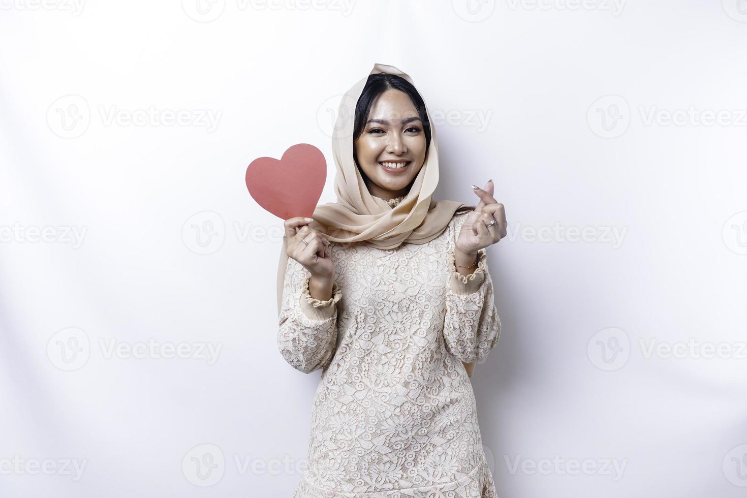 A happy young Asian Muslim woman wearing a hijab feels romantic shapes heart gesture expressing tender feelings and holding a red heart-shaped paper photo