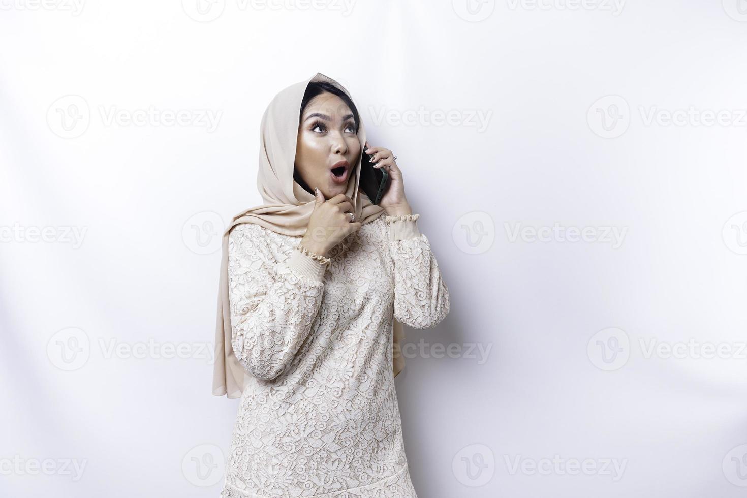 Shocked Asian woman wearing hijab, holding her phone, isolated by white background photo