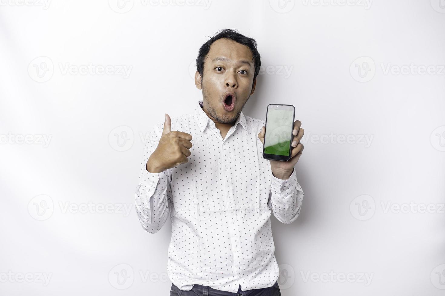 conmocionado asiático hombre vistiendo un blanco camisa y demostración verde pantalla en su teléfono, aislado por blanco antecedentes foto