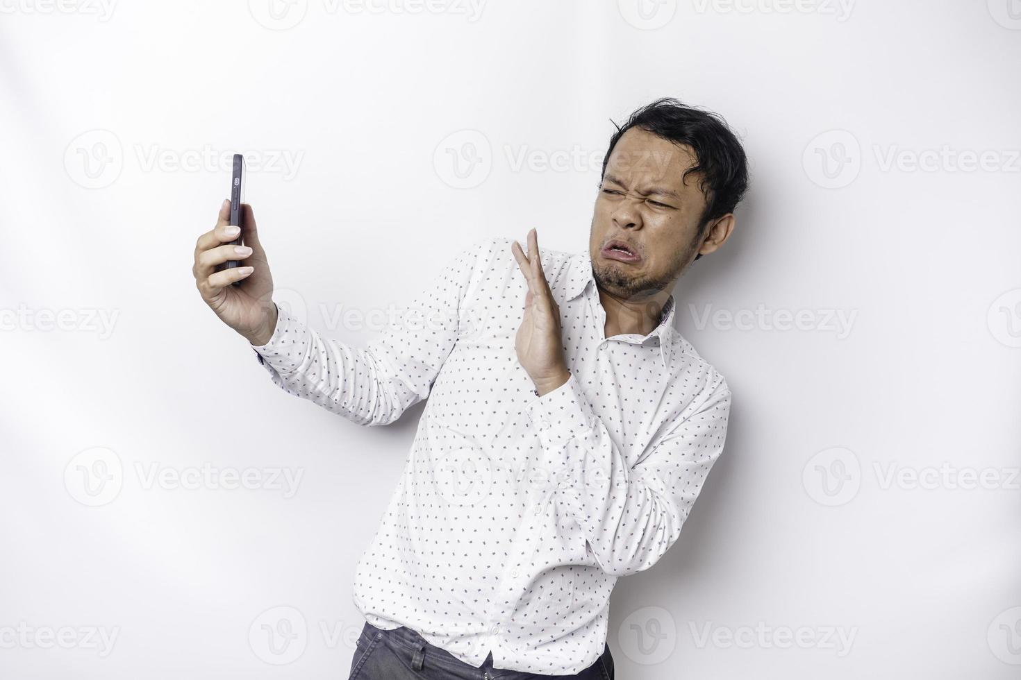 joven asiático hombre aislado en blanco fondo, mira deprimido, cara cubierto por brazos asustado y nervioso mientras participación su teléfono foto