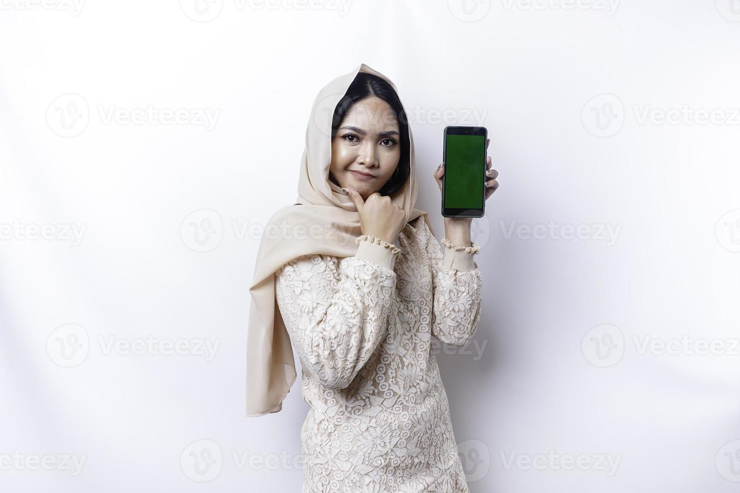 A thoughtful young Asian Muslim woman wearing a hijab and holding her chin while showing the phone screen isolated by a white background photo