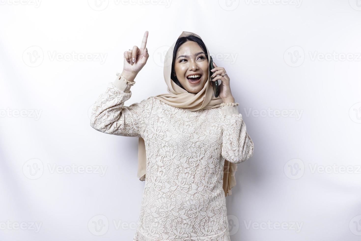 Excited Asian woman wearing hijab pointing at the copy space on top of her while holding her phone, isolated by white background photo