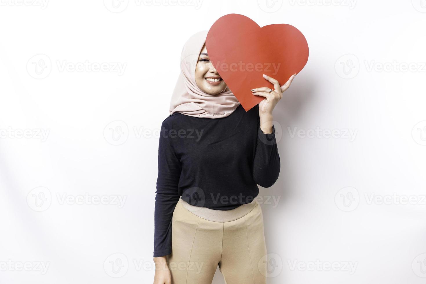 A happy young Asian Muslim woman wearing a hijab feels romantic shapes heart gesture expressing tender feelings and holding a red heart-shaped paper photo