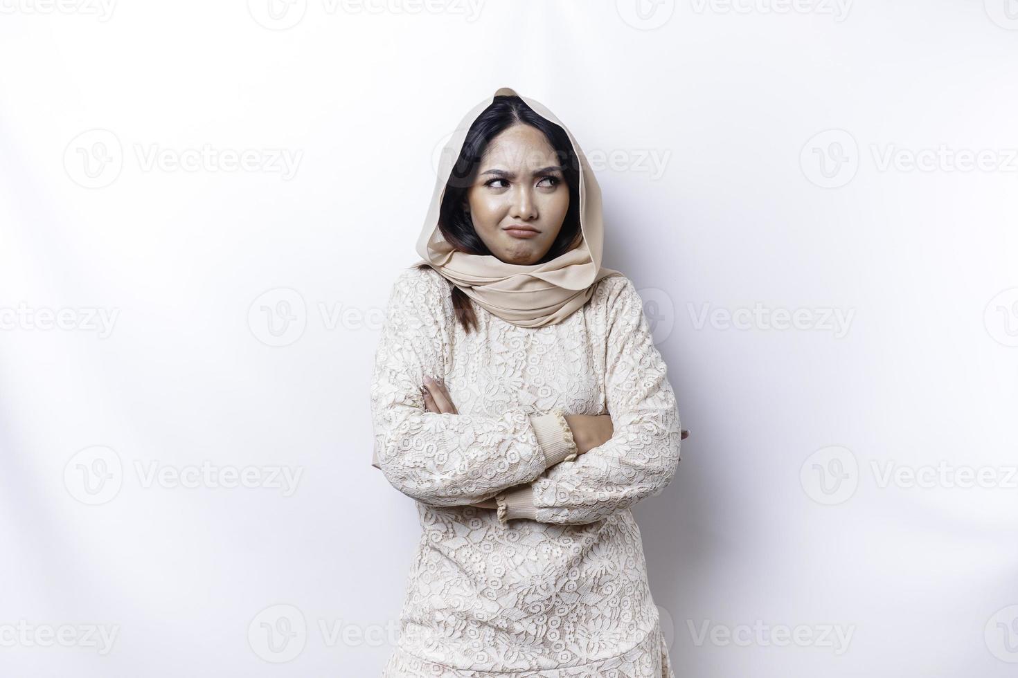 A dissatisfied young Asian Muslim woman looks at the camera posing on a white background, disgruntled girl with irritated face expressions show negative attitude photo