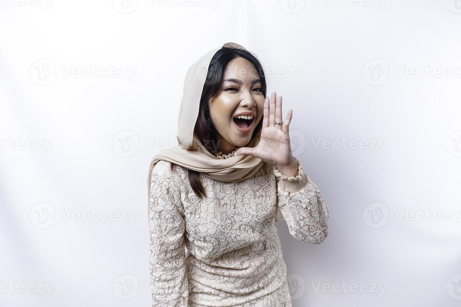 Young beautiful Asian Muslim woman wearing a hijab shouting and screaming loud with a hand on her mouth. communication concept. photo