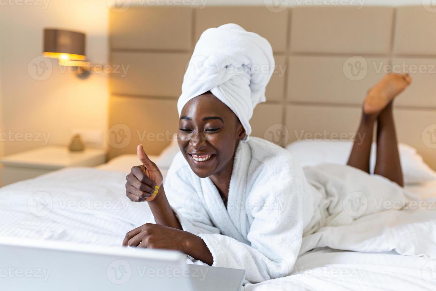 Beautiful young African woman in bathrobe resting on bed after shower while working on her laptop in the morning.Looking at laptop computer and talking to her friends via video call. photo