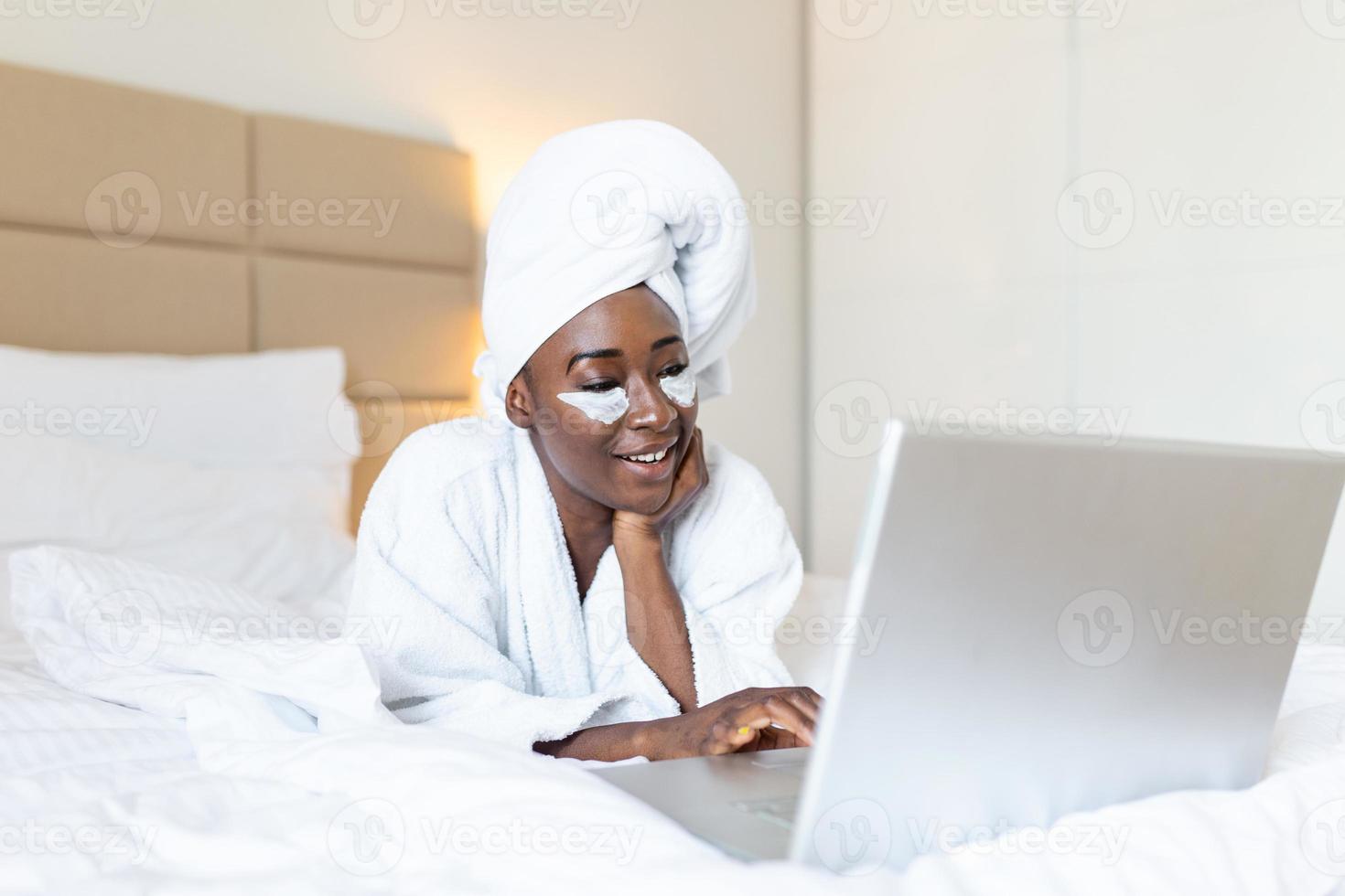 Smiling african young woman with face cream under her eyes lying on bed in bathrobe and towel on the head using a laptop on the bed at home photo