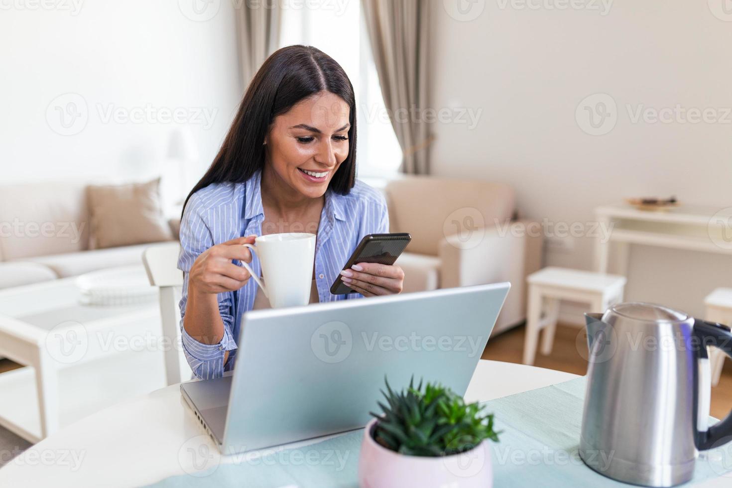 Self employed woman working with her phone and laptop at home with a cup of coffee. Happy woman using mobile phone while working at home with laptop. Smiling woman messaging with smartphone. photo