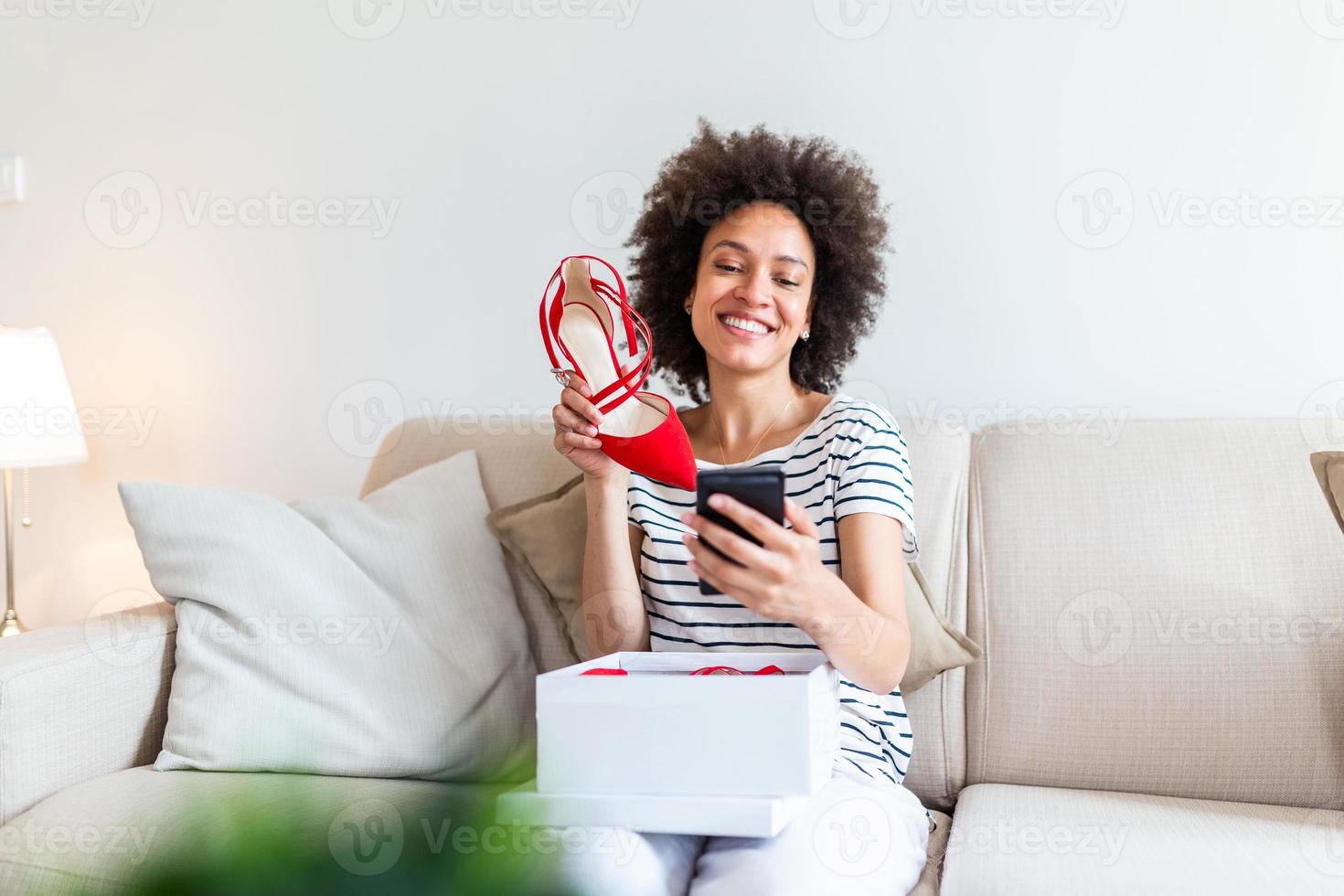Smiling woman unboxing a postal parcel and taking a selfie with her new purchases using a smartphone. Showing shoes to her friends via video call photo