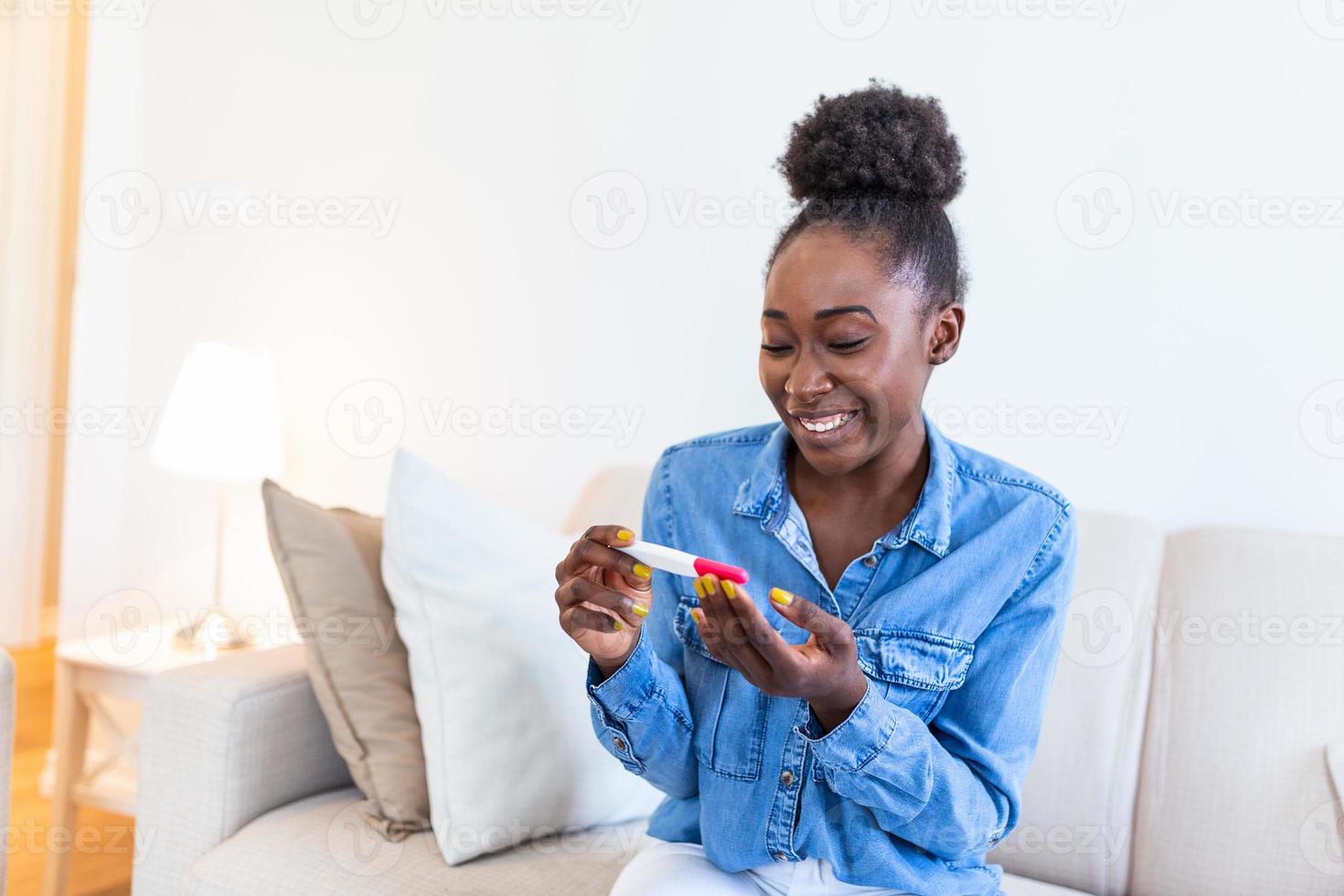 Young woman looking at pregnance test in happiness. Finally pregnant. Attractive black women looking at pregnancy test and smiling while sitting on the sofa at home photo