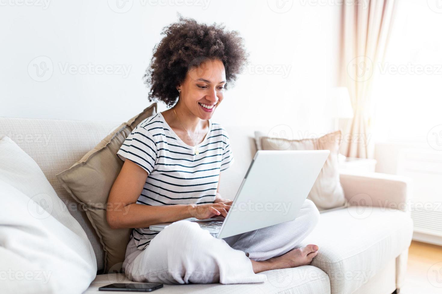 Young woman doing research work for her business. Smiling woman sitting on sofa relaxing while browsing online shopping website. Happy girl browsing through the internet during free time at home. photo