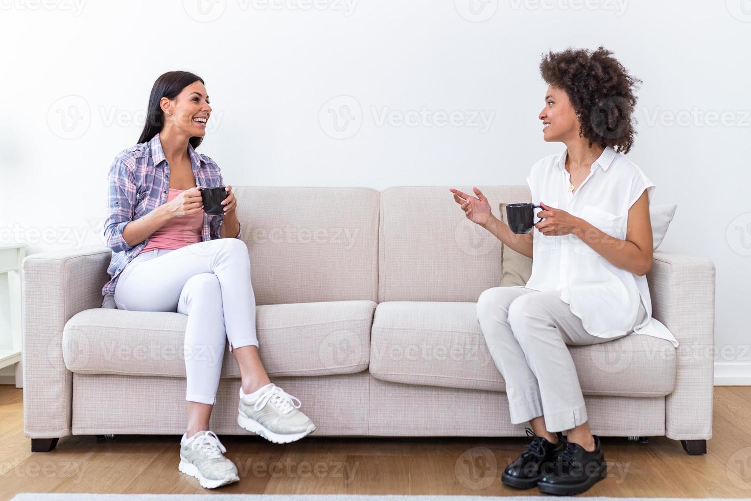 Two female friends in social distancing sitting on sofa. Best friends having coffee together while separated by social distancing on sofa at home photo