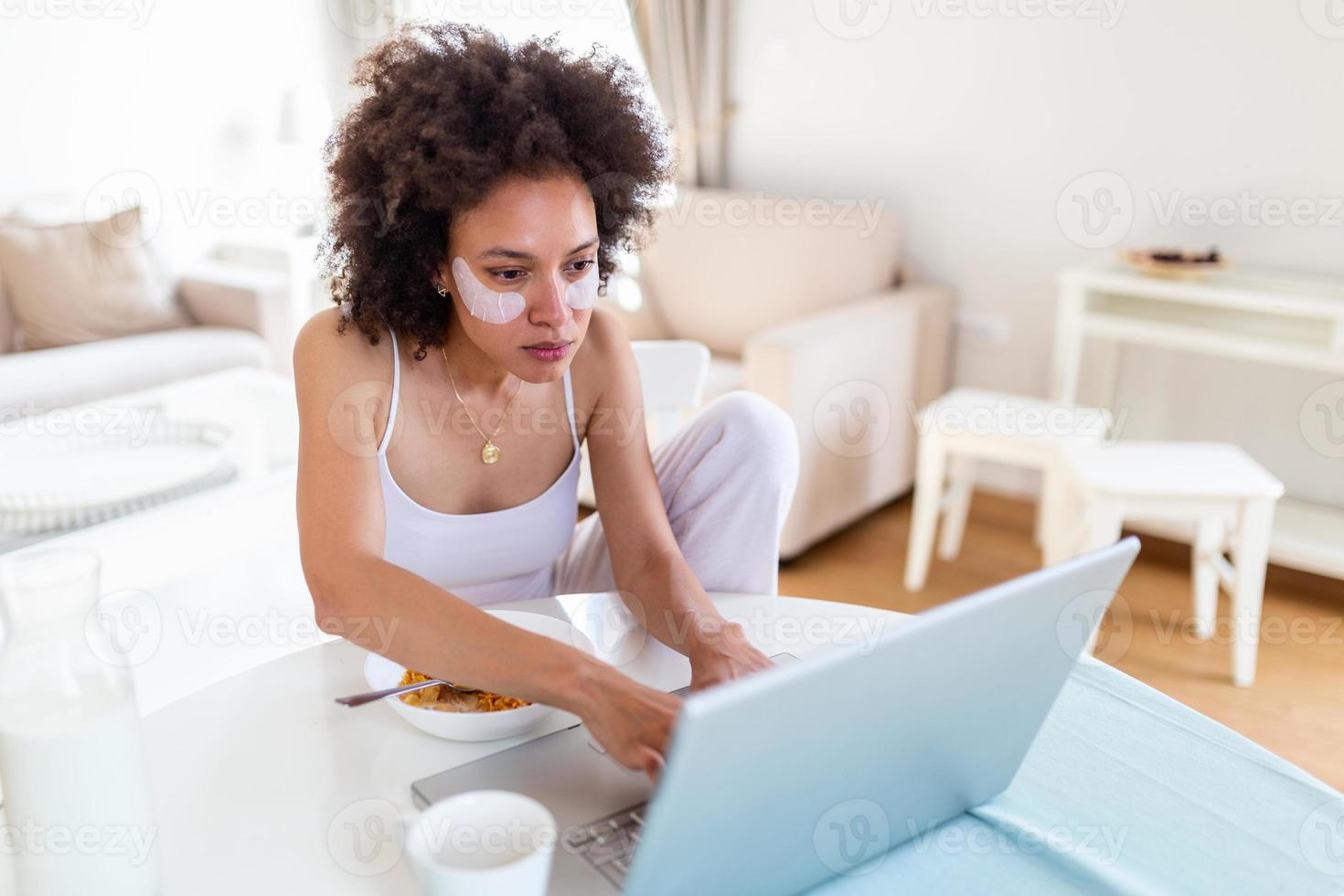 Beautiful young arfican american woman typing on laptop computer at home. Cheerful lady eating corn flakes and working on laptop at home photo