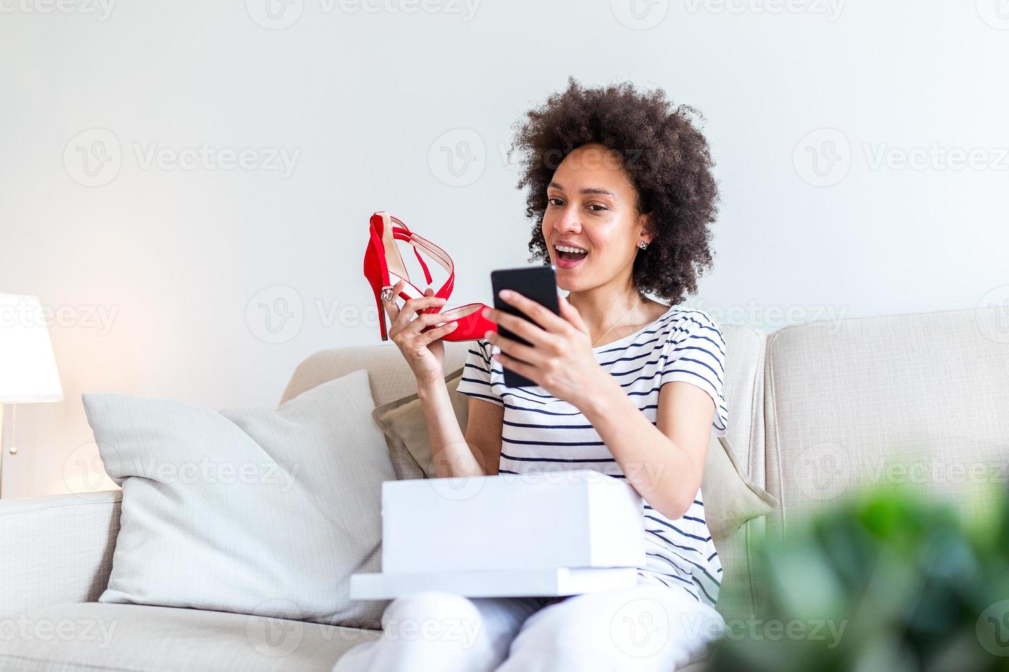 Smiling woman unboxing a postal parcel and taking a selfie with her new purchases using a smartphone. Showing shoes to her friends via video call photo