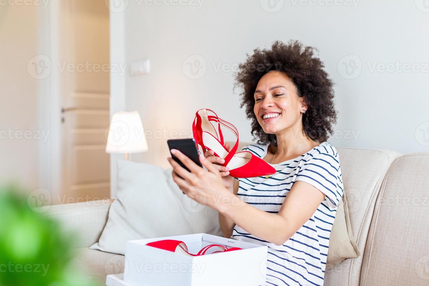 Smiling woman unboxing a postal parcel and taking a selfie with her new purchases using a smartphone. Showing shoes to her friends via video call photo