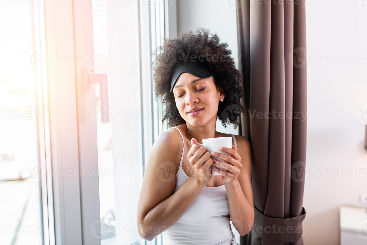Woman in the morning. Attractive woman with sleeping mask is holding a cup with hot tea or coffee and looking at the sunrise standing near the window in her home and having a perfect cozy morning. photo