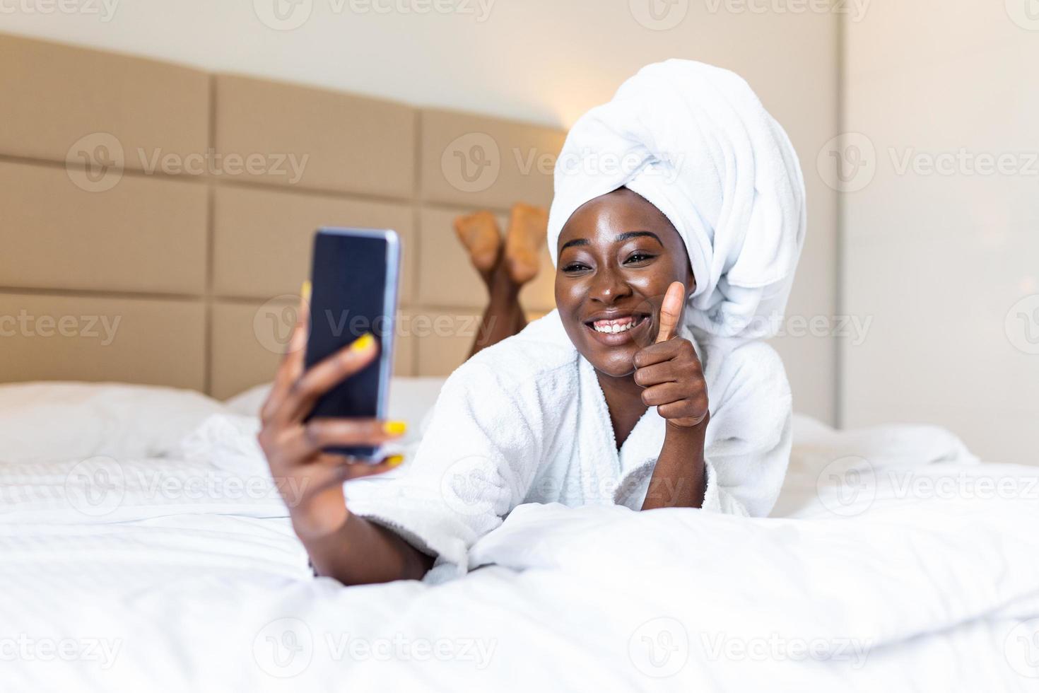 Smiling african woman lying on bed in bathrobe with mobile phone taking