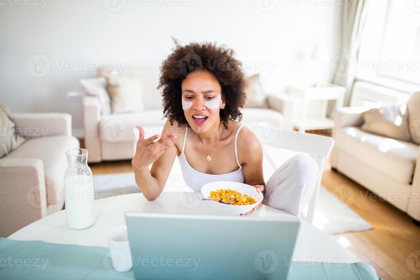 Image of happy young amazing woman sitting indoors at the table with laptop holding corn flakes. Looking at laptop computer and talking to her friends via video call. photo