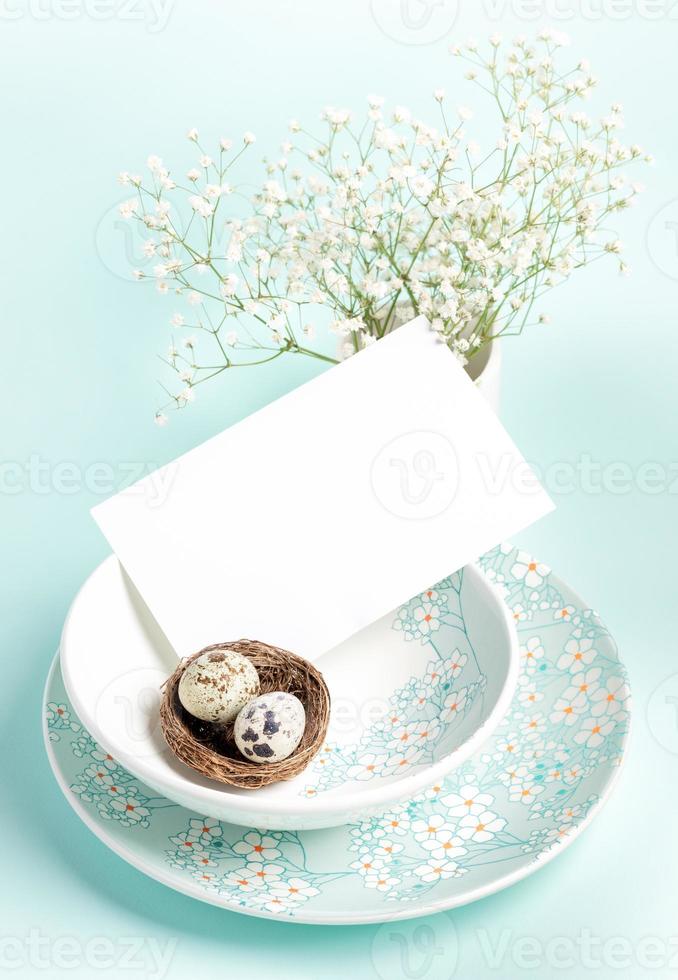 Easter card. Two quail eggs in nest, white empty note on plates with floral pattern on light blue. photo