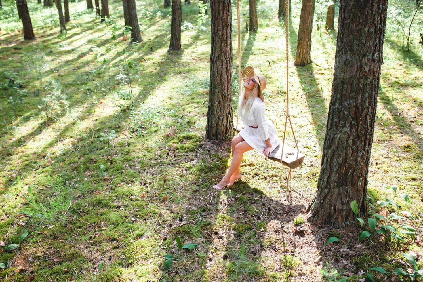 Blond young woman with slender naked legs in white dress and straw hat sitting on a rope swing photo