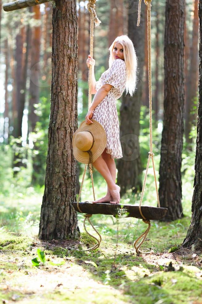 Blond young woman with slender naked legs in short dress and straw hat posing on a rope swing photo