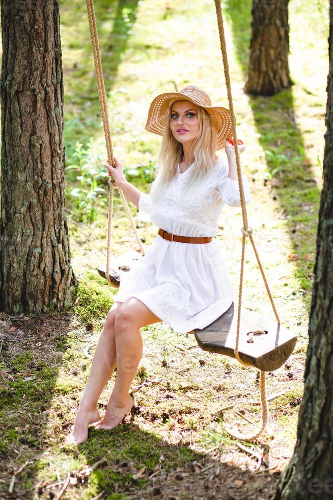 Blond young woman with slender naked legs in white dress and straw hat sitting on a rope swing photo