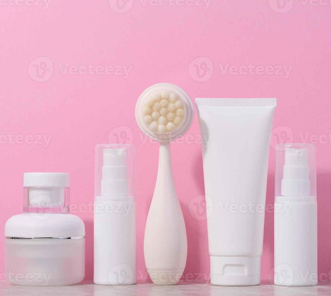 White plastic tubes and jars of cream, and a massage brush for facial cleansing on a pink background, items for cosmetic procedures photo