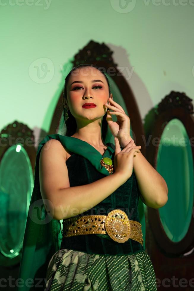 a female Javanese dancer in a green dance costume and a full face with makeup poses with her hands in front of a dressing table photo