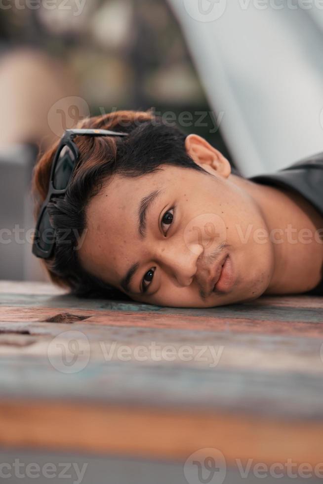 un asiático hombre con un gordito cara vistiendo Gafas de sol y un negro cuero chaqueta mientras sentado a un café mesa foto