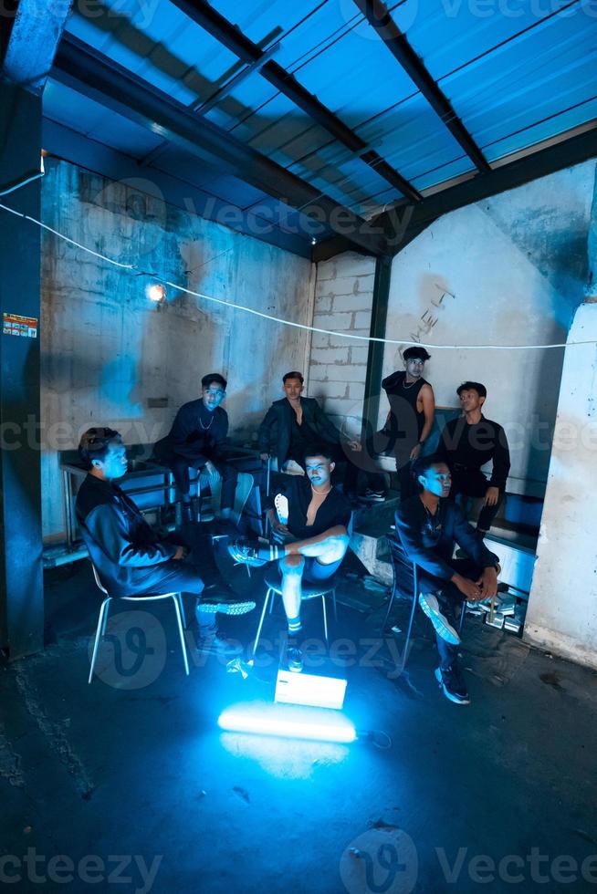 un grupo de asiático adolescentes en negro ropa sentado juntos en un negro edificio con un azul ligero en el habitación foto