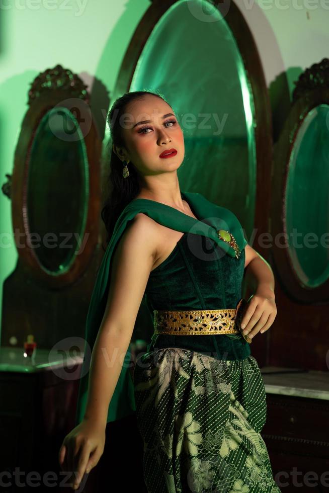 un artista mujer con un verde vestir y bufanda posando muy hermosa y exótico en frente de un maquillaje espejo en un verde habitación foto