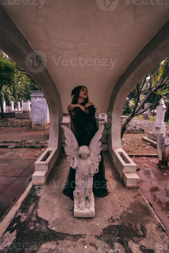 a young witch in a black dress is standing between the tombstones shaped like an angel while visiting the cemetery photo