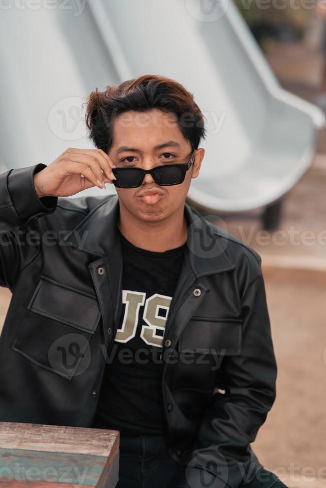 un asiático hombre es sentado en un de madera silla mientras vistiendo un negro cuero camisa y chaqueta mientras elegante con Gafas de sol foto