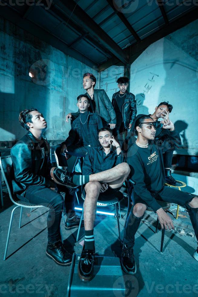 a group of Asian teenagers in black clothes posing very coolly while sitting in a warehouse chair which is lit blue photo