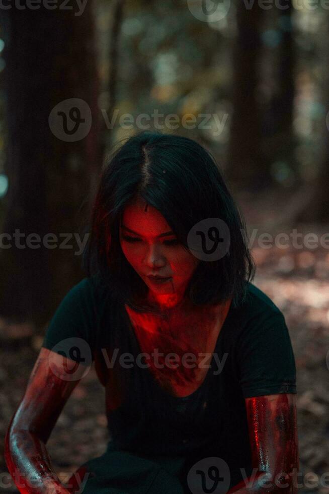 a female serial killer in black clothes and covered in blood has a scary face sitting in the middle of a forest photo