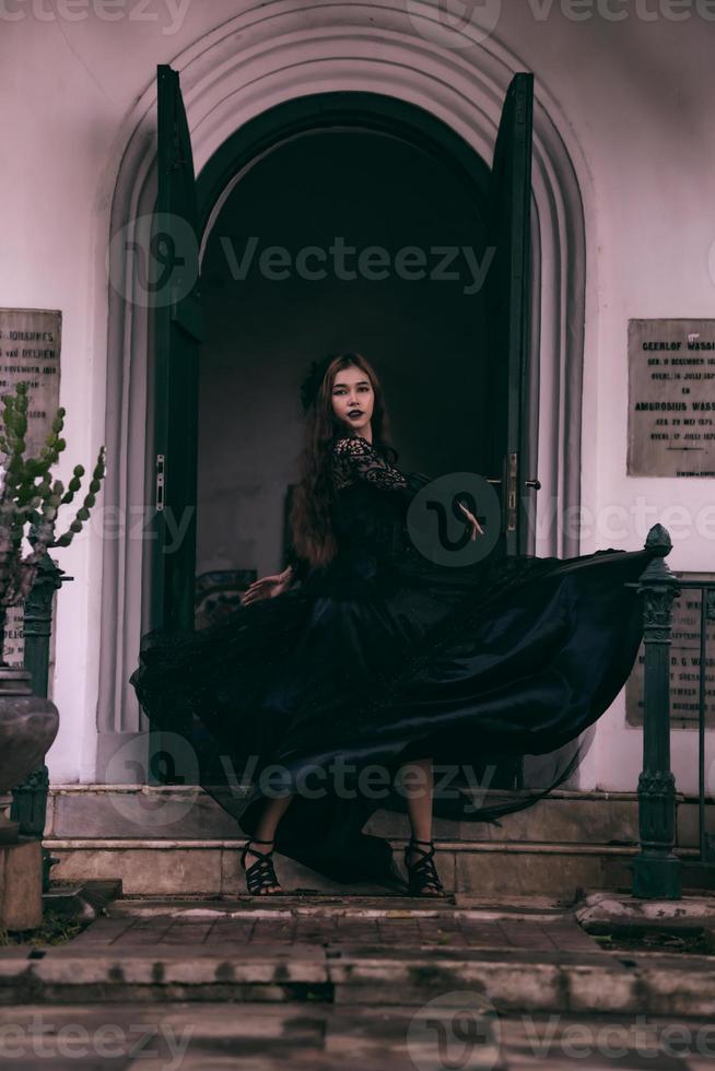 an Asian teenager posing in her black dress floating in the air while standing in front of a cemetery with all black makeup on her face photo