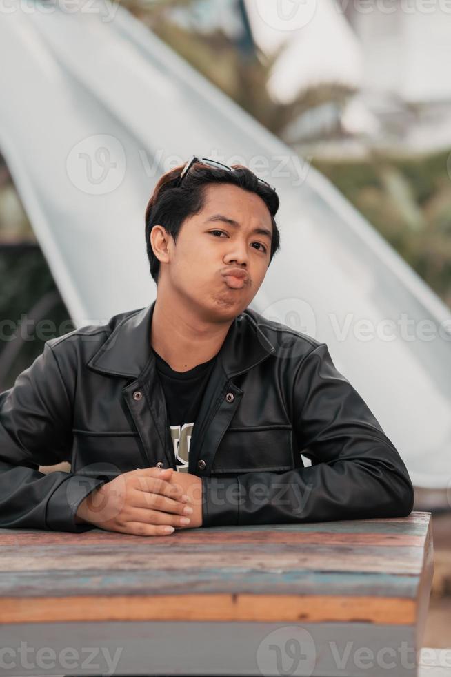 an Asian man with a chubby face wearing sunglasses and a black leather jacket while sitting at a cafe table photo