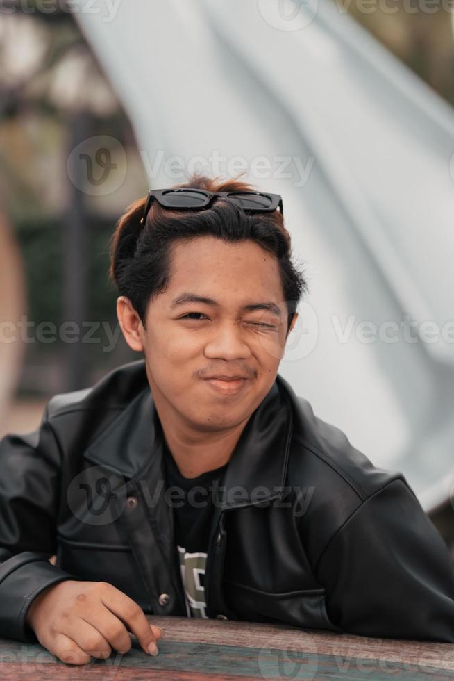 an Asian man with a chubby face wearing sunglasses and a black leather jacket while sitting at a cafe table photo