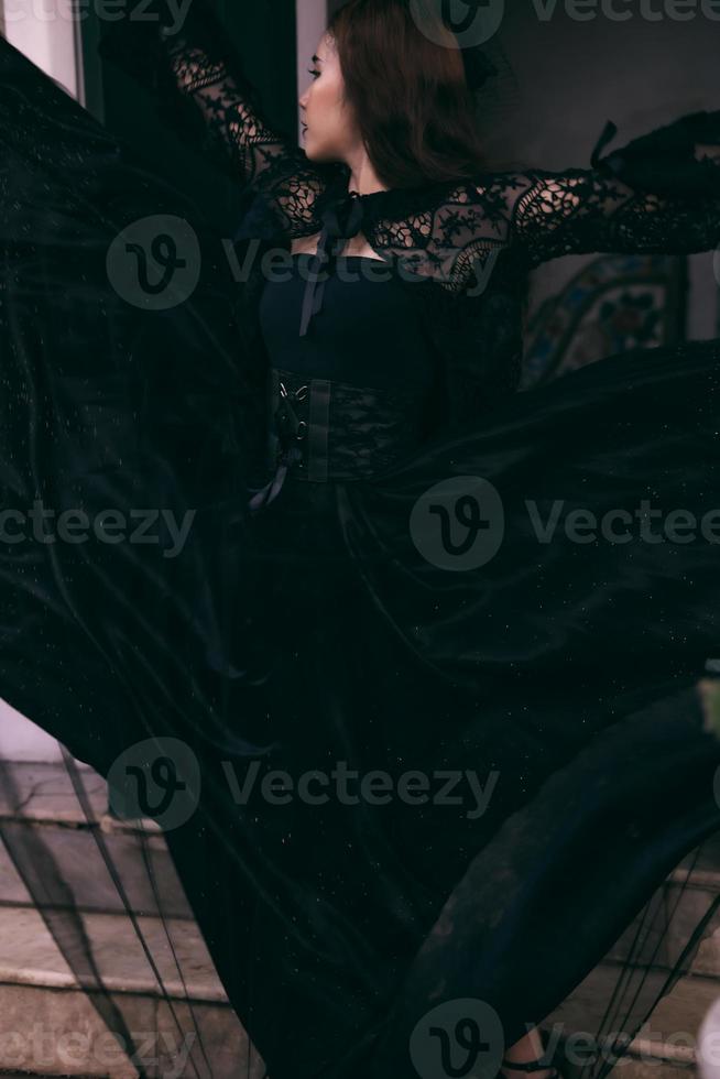 an Asian woman with a witch-like appearance flung her all-black skirt in the air as she stood in front of a cemetery photo