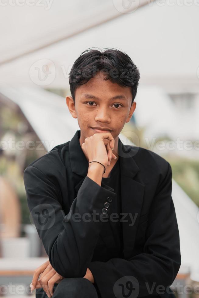 un asiático adolescente en un negro mezclilla chaqueta se inclina en contra un jardín mesa mientras relajante y disfrutando el ver foto